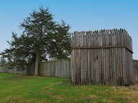 Wood stockade twice the height of a person with evergreen tree beside.