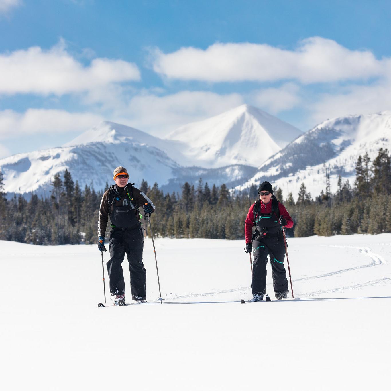 Two skiers travel across a flat meadow with mountains in the distance.