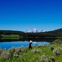 A hiker walks along the lakeshore with mountains in the background.