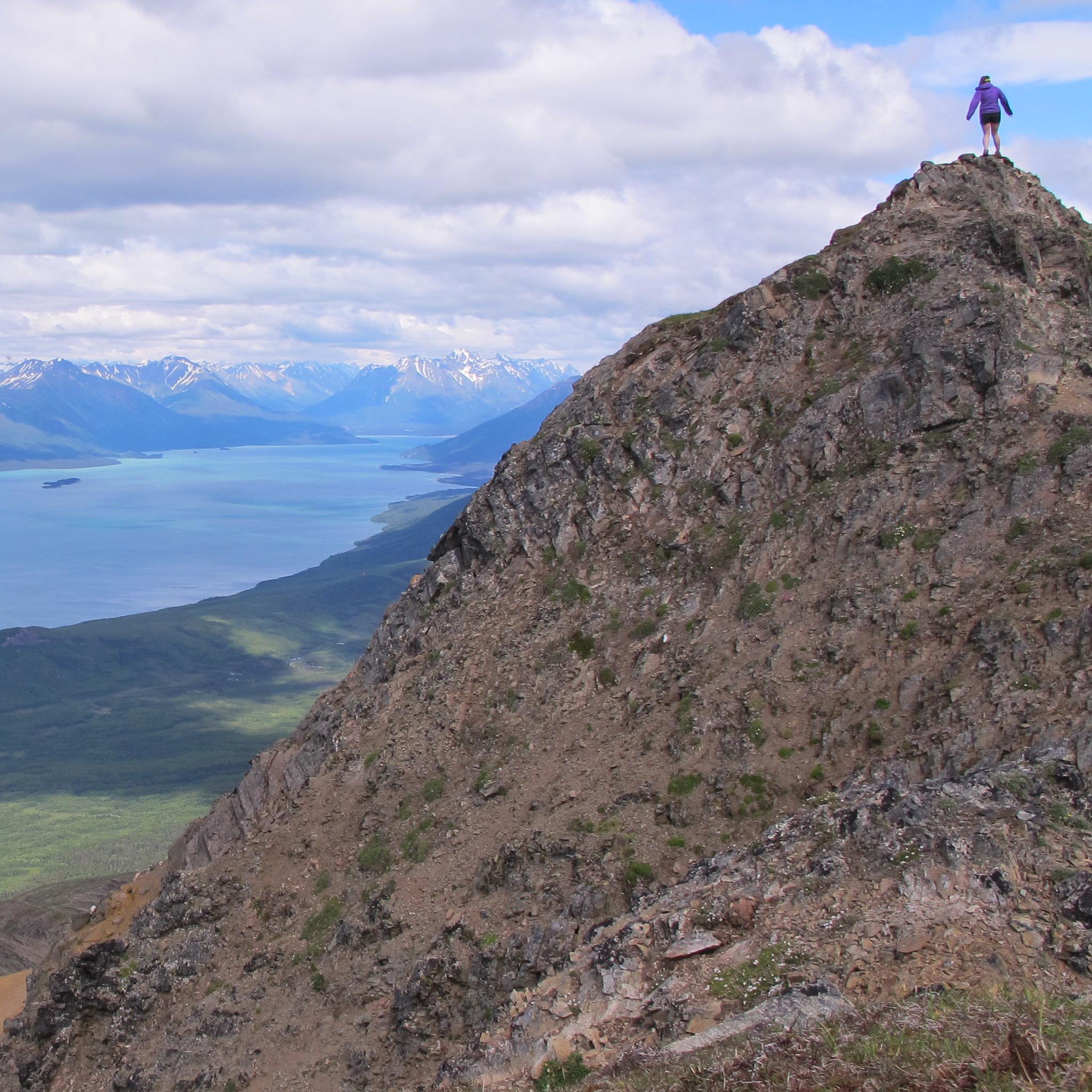 A woman stands on top of a rocky mountain looking out towards the turquoise water of Lake Clark