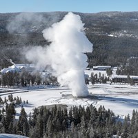 A geyser in the middle of a snowy field erupts steam and water high into the air.