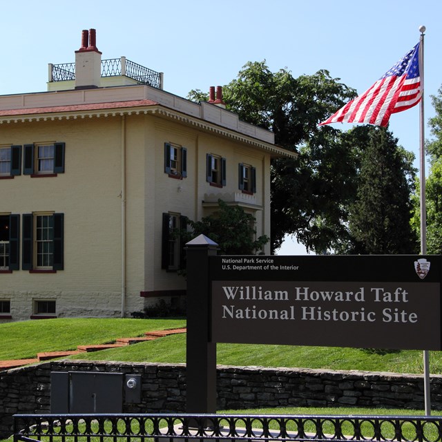 The large Taft house in the background and a park sign in foreground with U.S. flag flying between