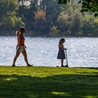 A color photo of a woman and girl on a scooter along a path that is beside water.