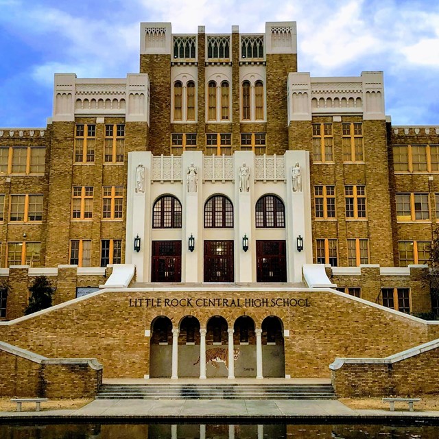 A large brown brick school with a reflecting pond and staircases in front. 