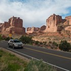 a tan car drives on a road with rock walls and pinnacles in the background