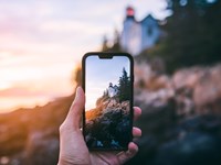 A hand holds a phone poised to snap a photo of a lighthouse bathed in sunset on a rocky coastline.