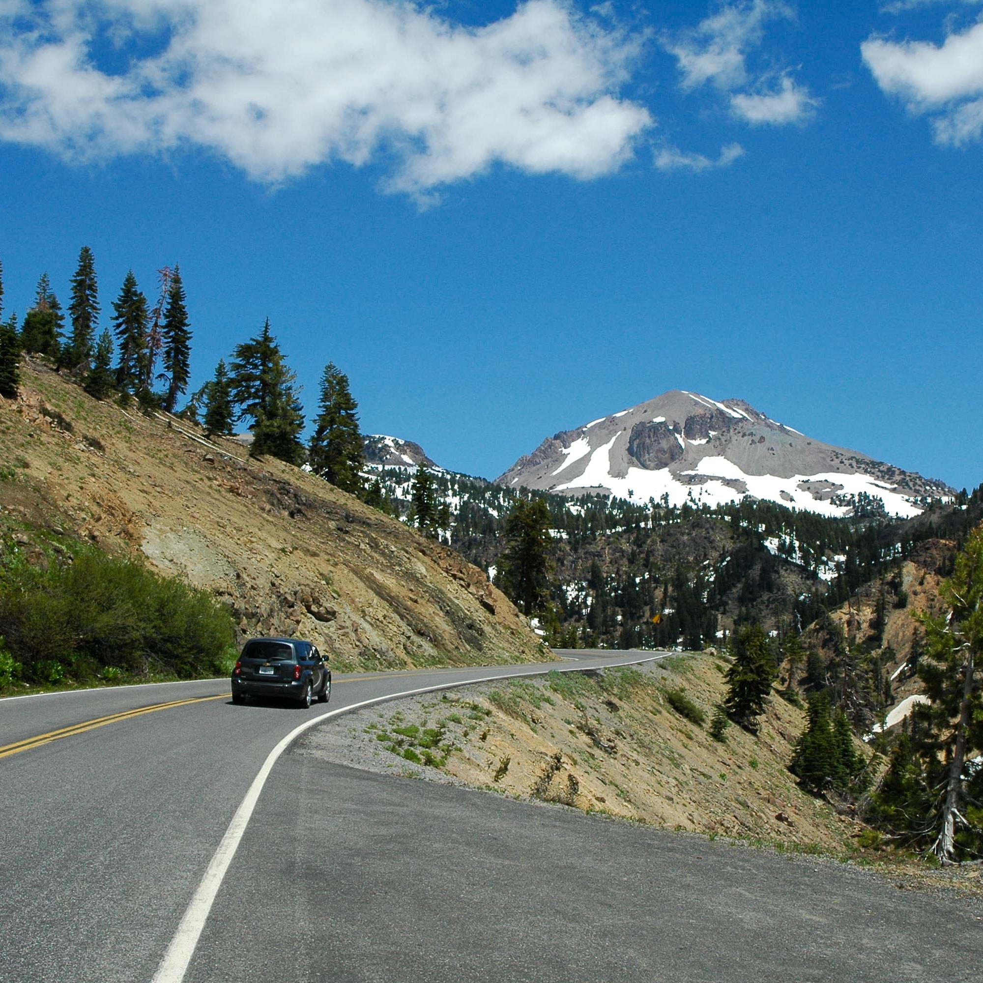 A black vehicle drives on a mountain road toward a large volcanic peak with large patches of snow.