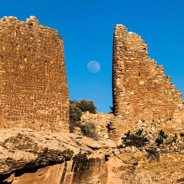 two stone structures on a cliff edge. The full moon rises between them