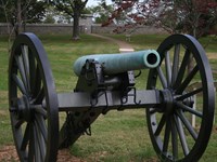 A green colored cannon faces the viewer. a cube shaped monument behind a stone wall in the distance.
