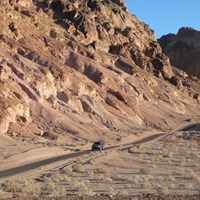 A vehicle drives along a single lane road through colorful, eroded hills. 