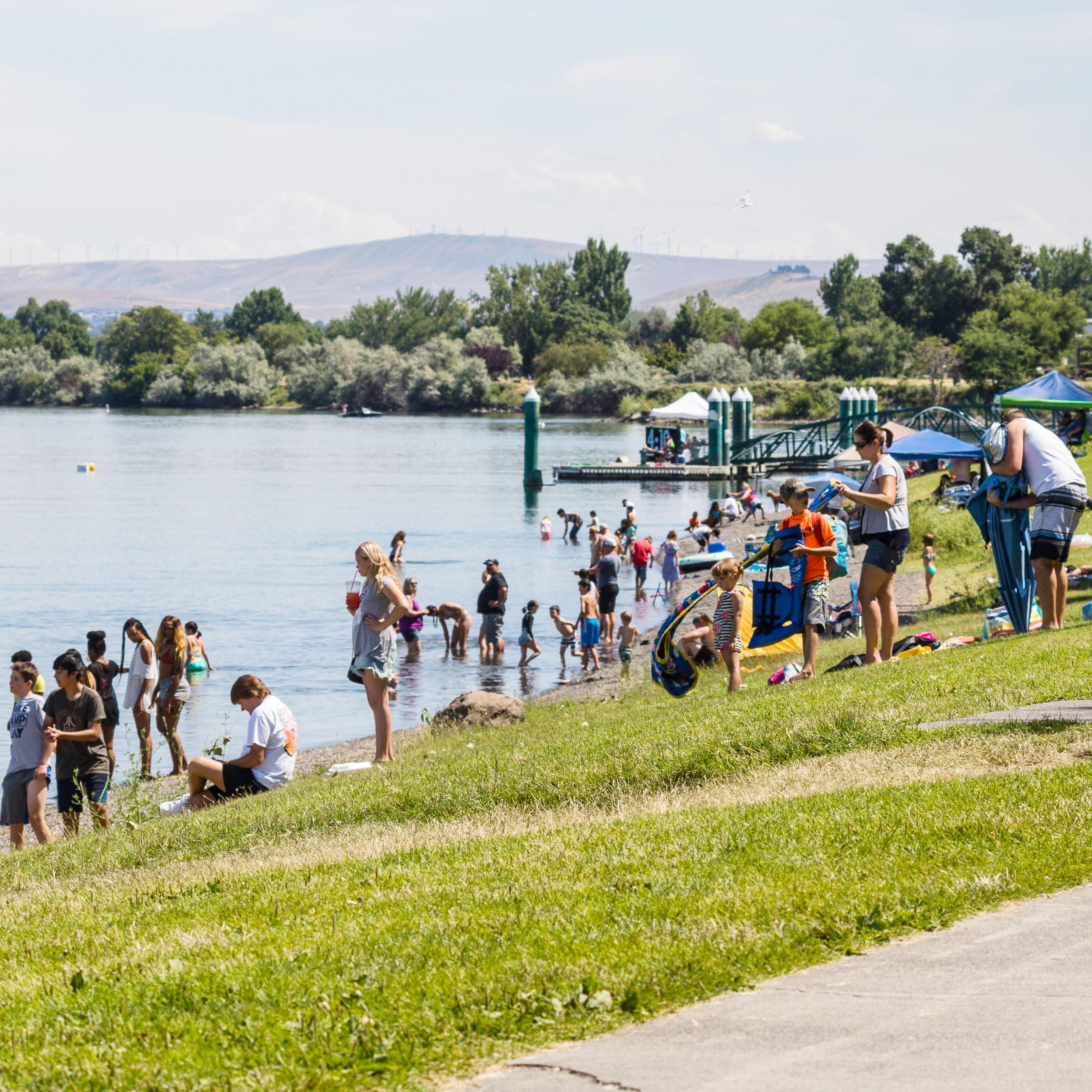 Color photograph of a river\'s shore with numerous people gathered on a green lawn beside it.