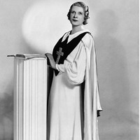 Black and white photograph of Aimee Semple McPherson standing behind a white podium.