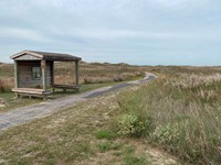 A paved trail leads into a wide-open prairie. A small shelter with benches is next to the trail.