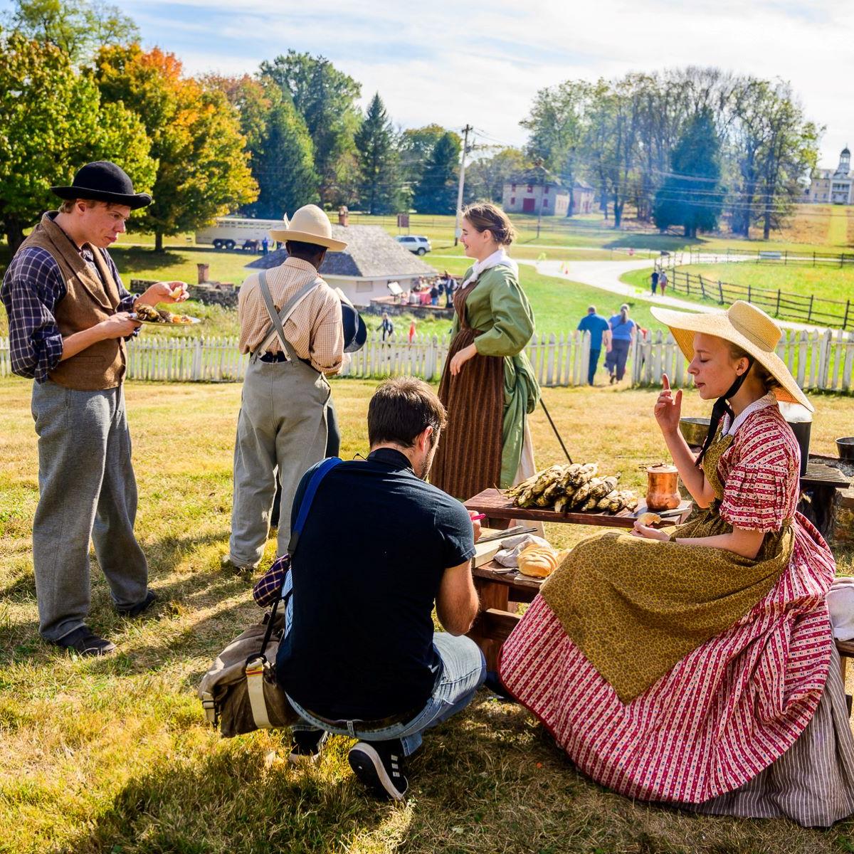 Living historians eating food at a wooden table on a sunny day.