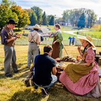 Living historians eating food at a wooden table on a sunny day.