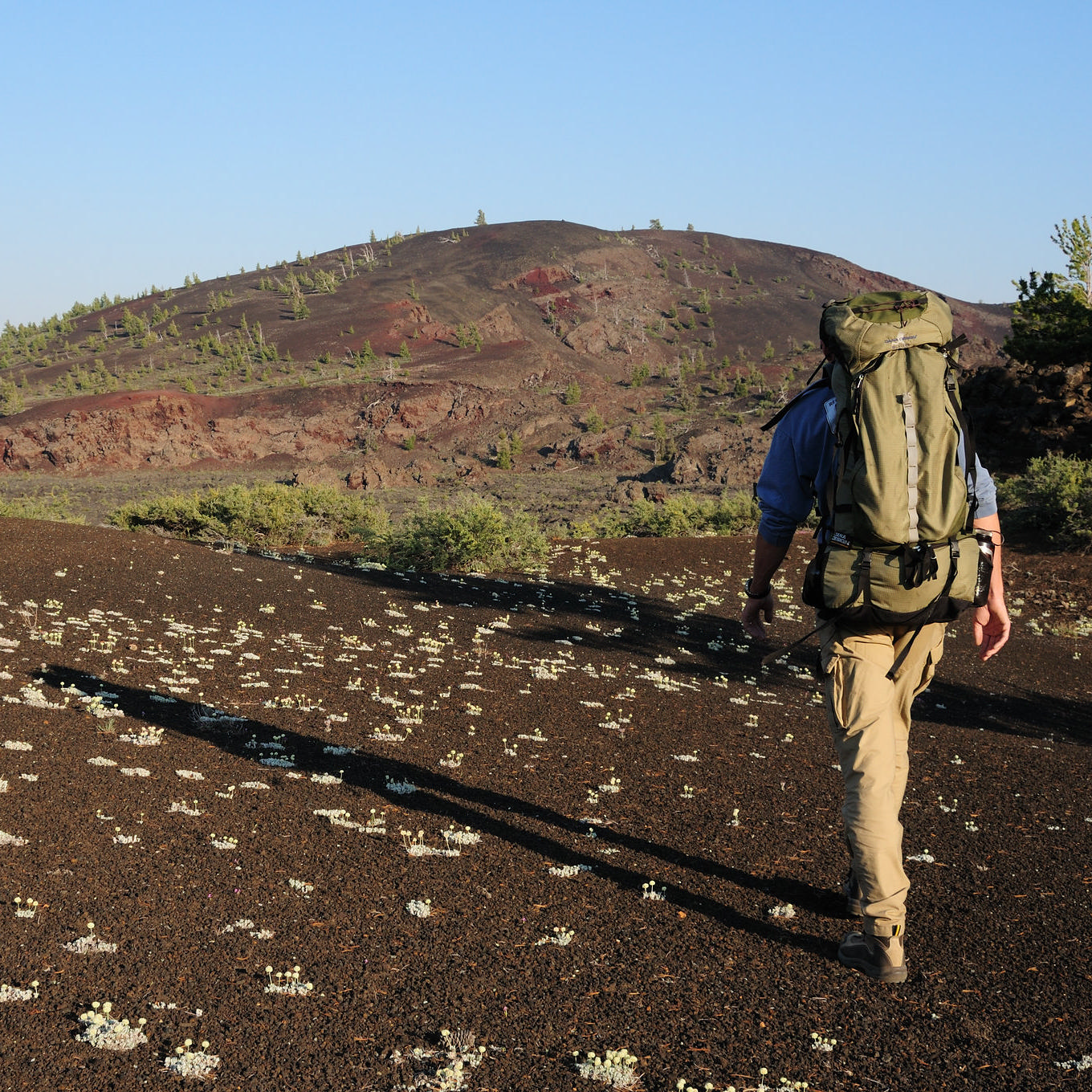 A hiker with a large backpack walks on a cinder trail toward a large volcanic cone.