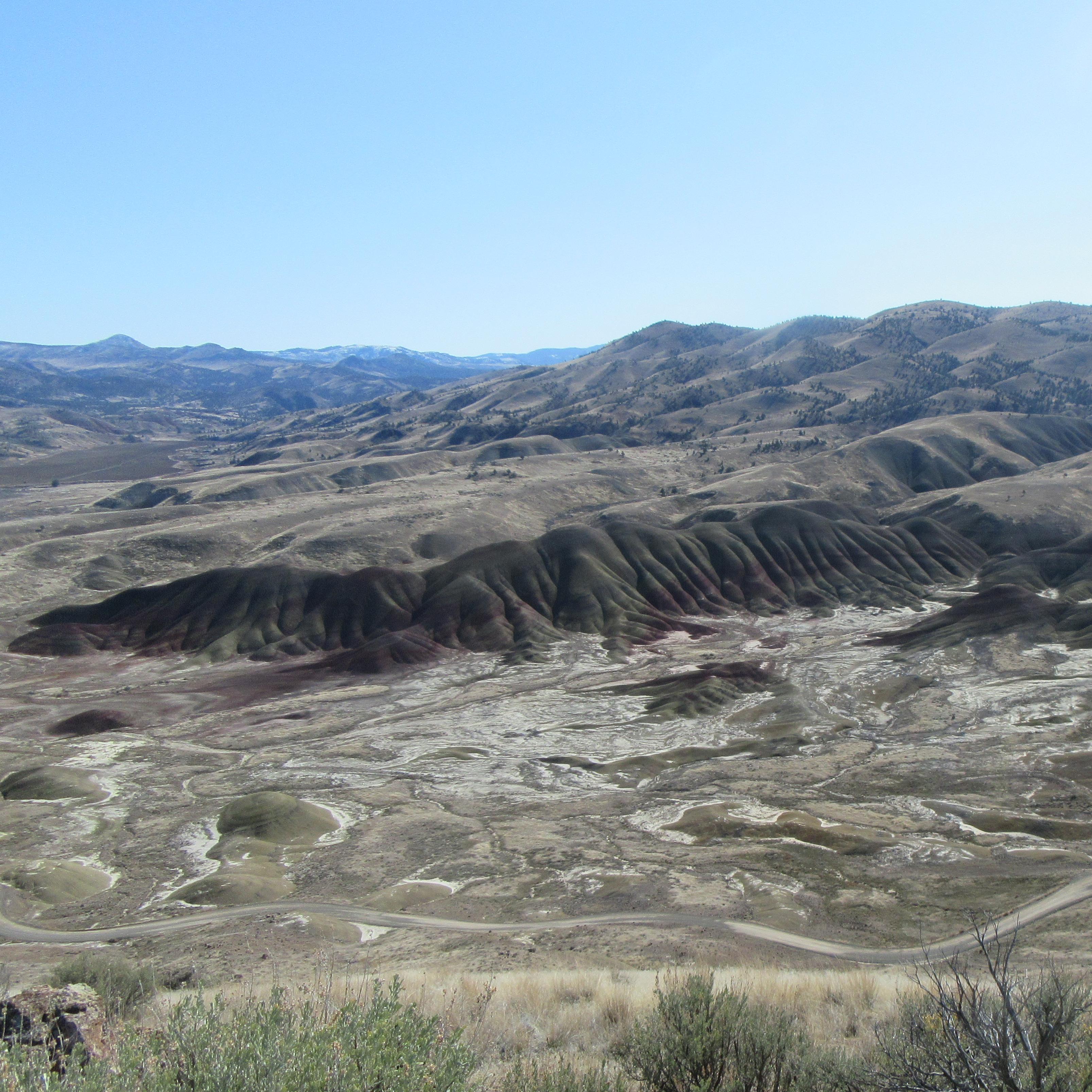 View of red and green striped hills from trail above, with green sagebrush and windy dirt road below