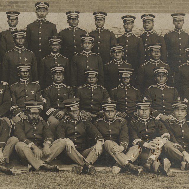 Black soldiers sitting and standing in dark-colored uniforms and caps posing for a group photo
