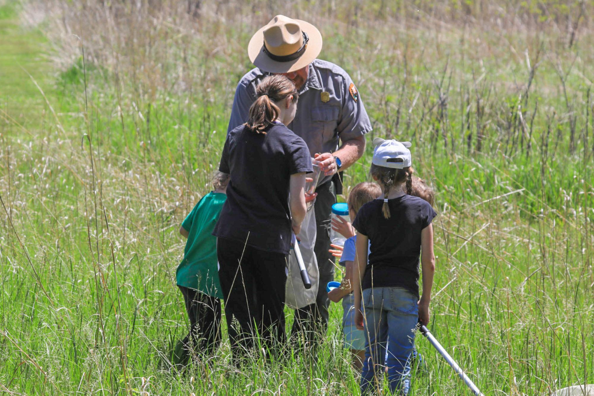 Five youth with bug nets and clear jars surround a uniformed ranger on the prairie.