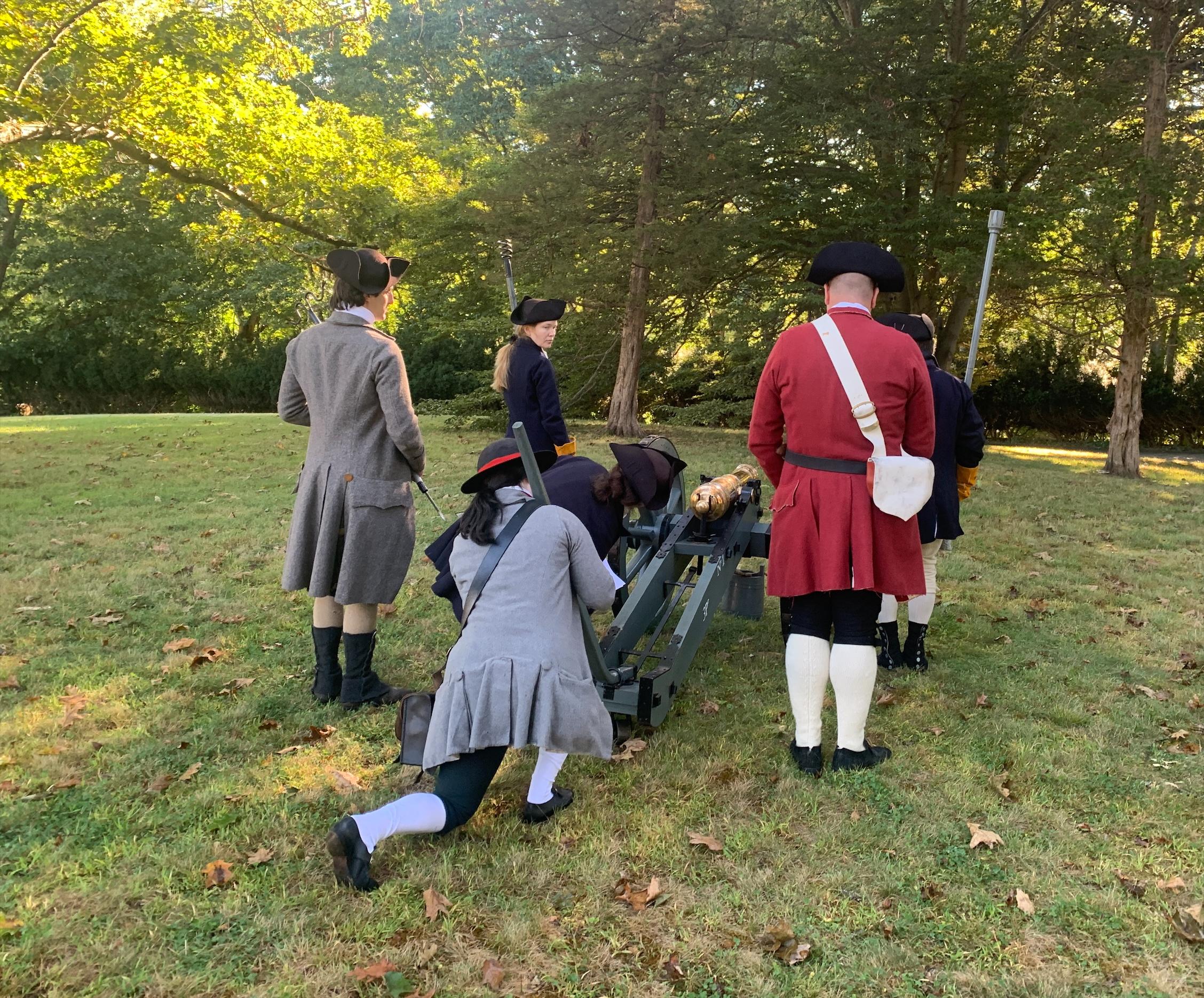 A crew of soldiers in 18th century clothing stand around a cannon to take aim