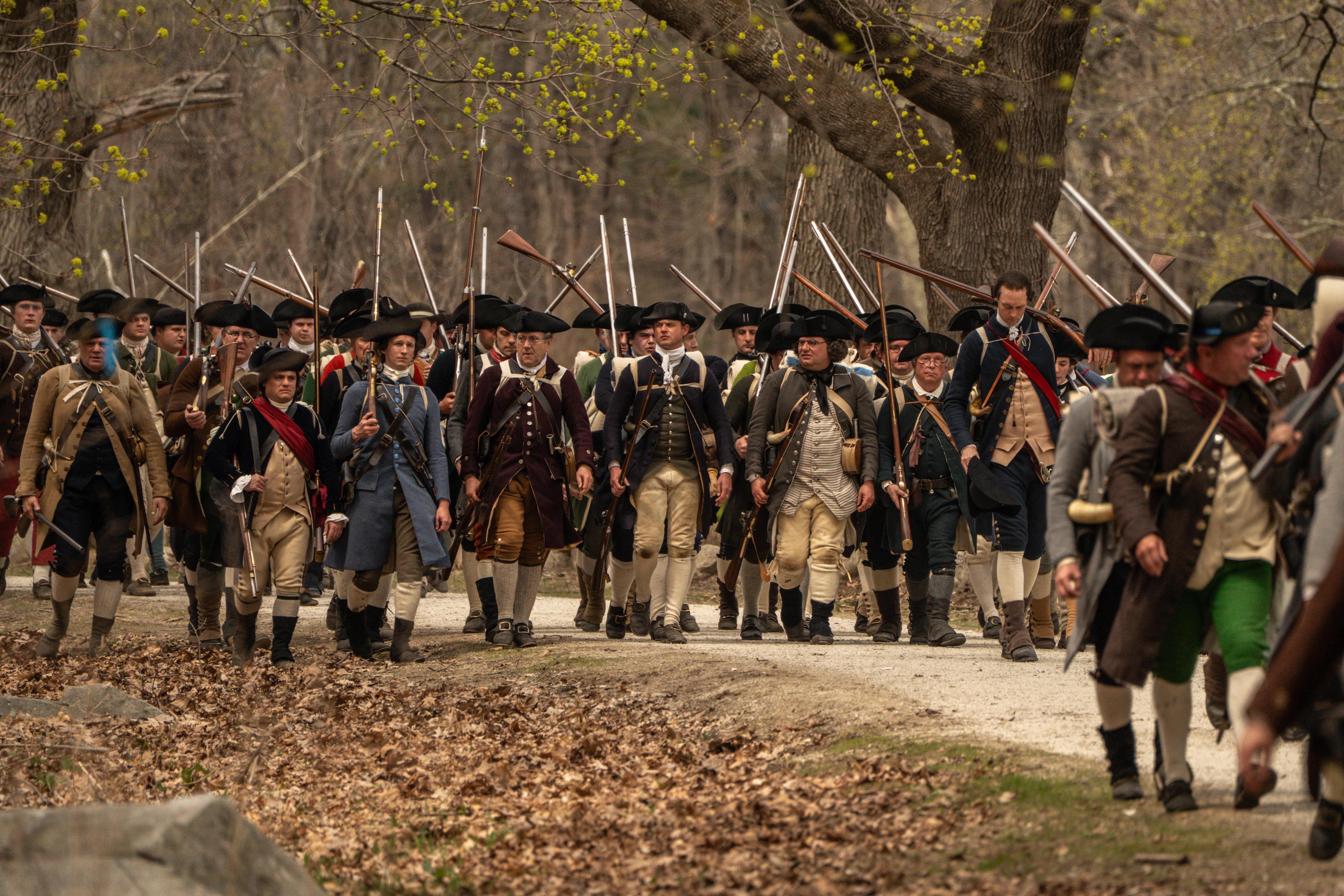 A large group of militia soldiers march down a country road. They carry muskets and military gear