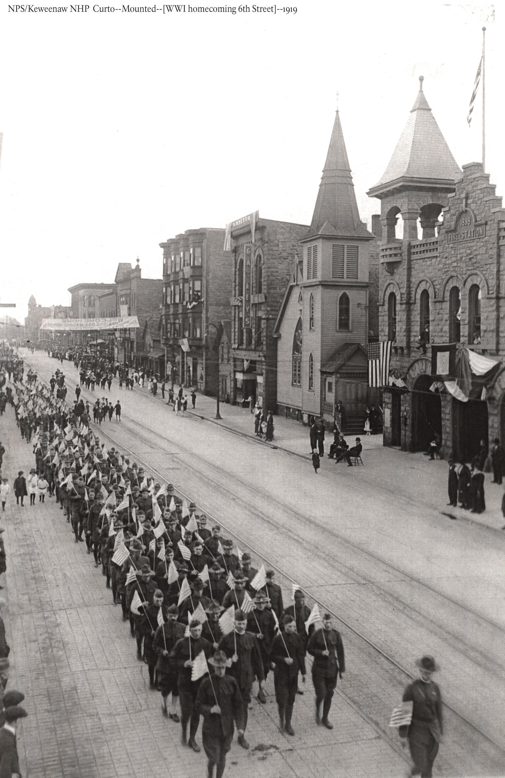 Uniformed military members march down a village street with buildings on each side