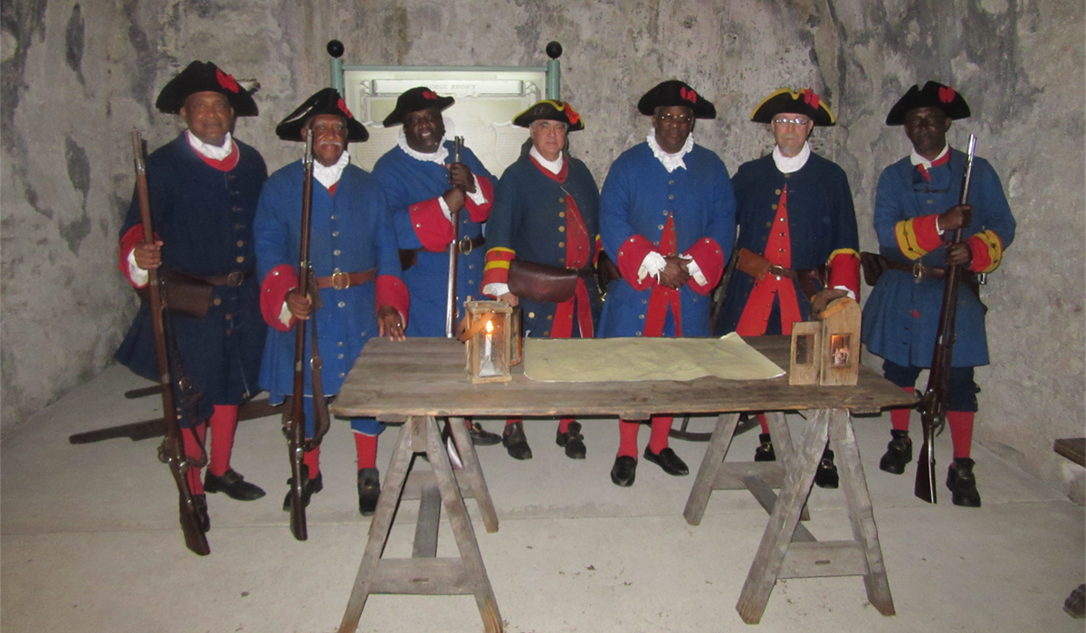 7 men in 1740 Spanish uniforms, 4 of them holding muskets, behind a wooden table set with lanterns