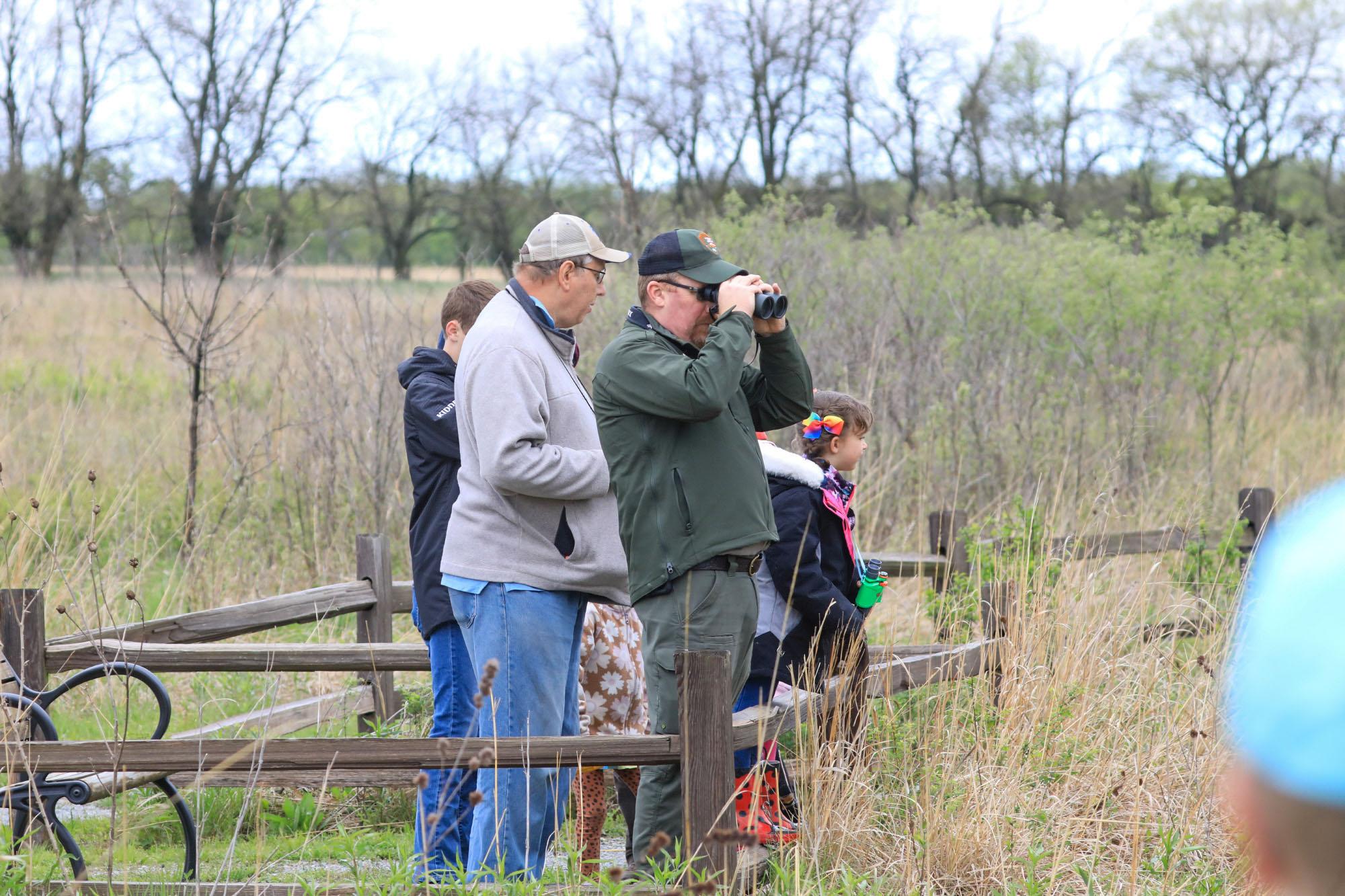 Outdoor uniformed park ranger looks through binoculars and talks to nearby visitors.