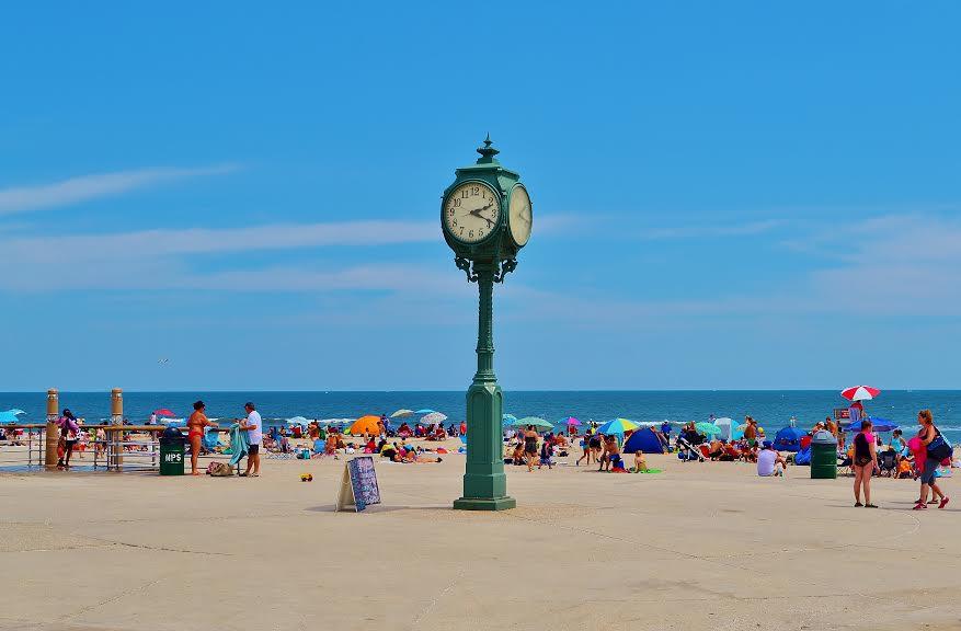 Green clock on boardwalk at beach with people, umbrellas and chairs behind it