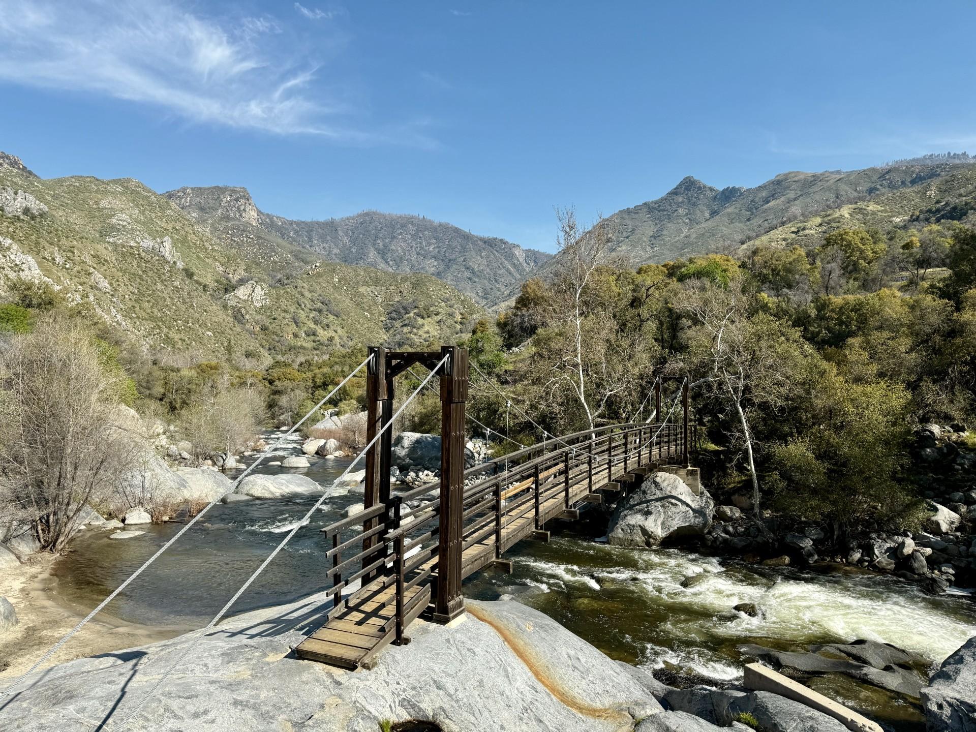 A narrow wooden suspension bridge crosses a rushing river with mountains and trees in the background