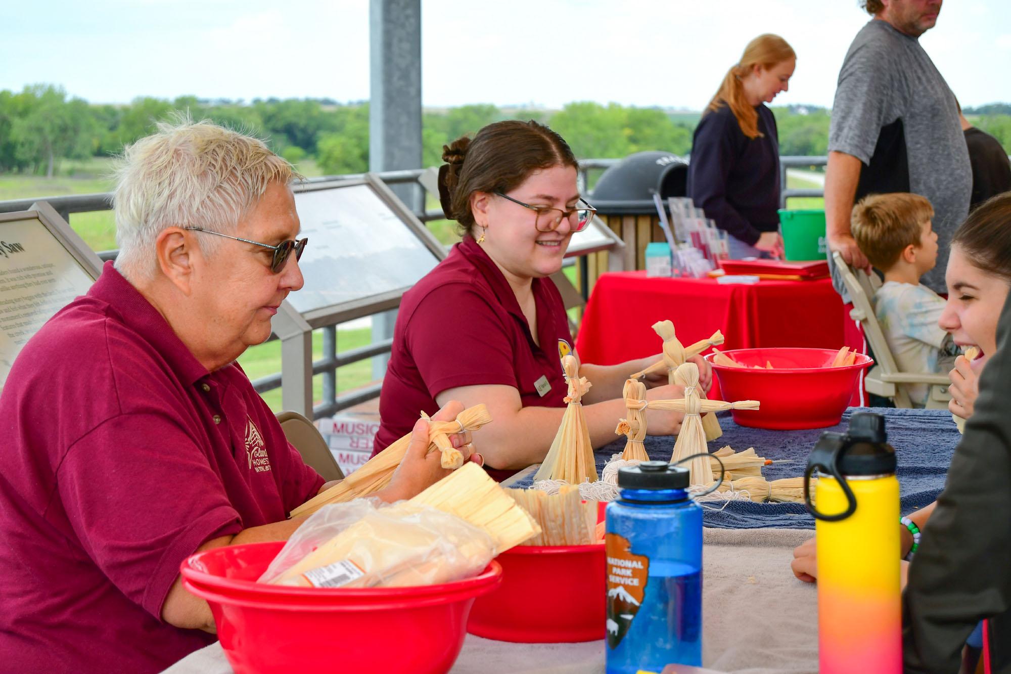 Two Volunteers in Parks assist park visitors in making corn husk dolls.