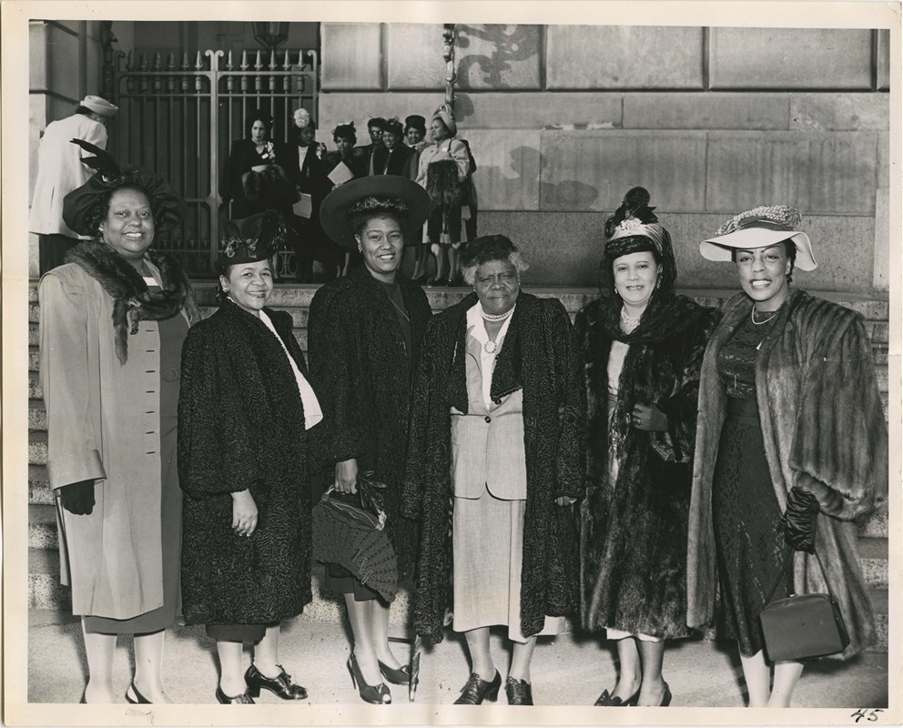 Six woman stand outside the front a building wearing hats, gloves, and fur coats.