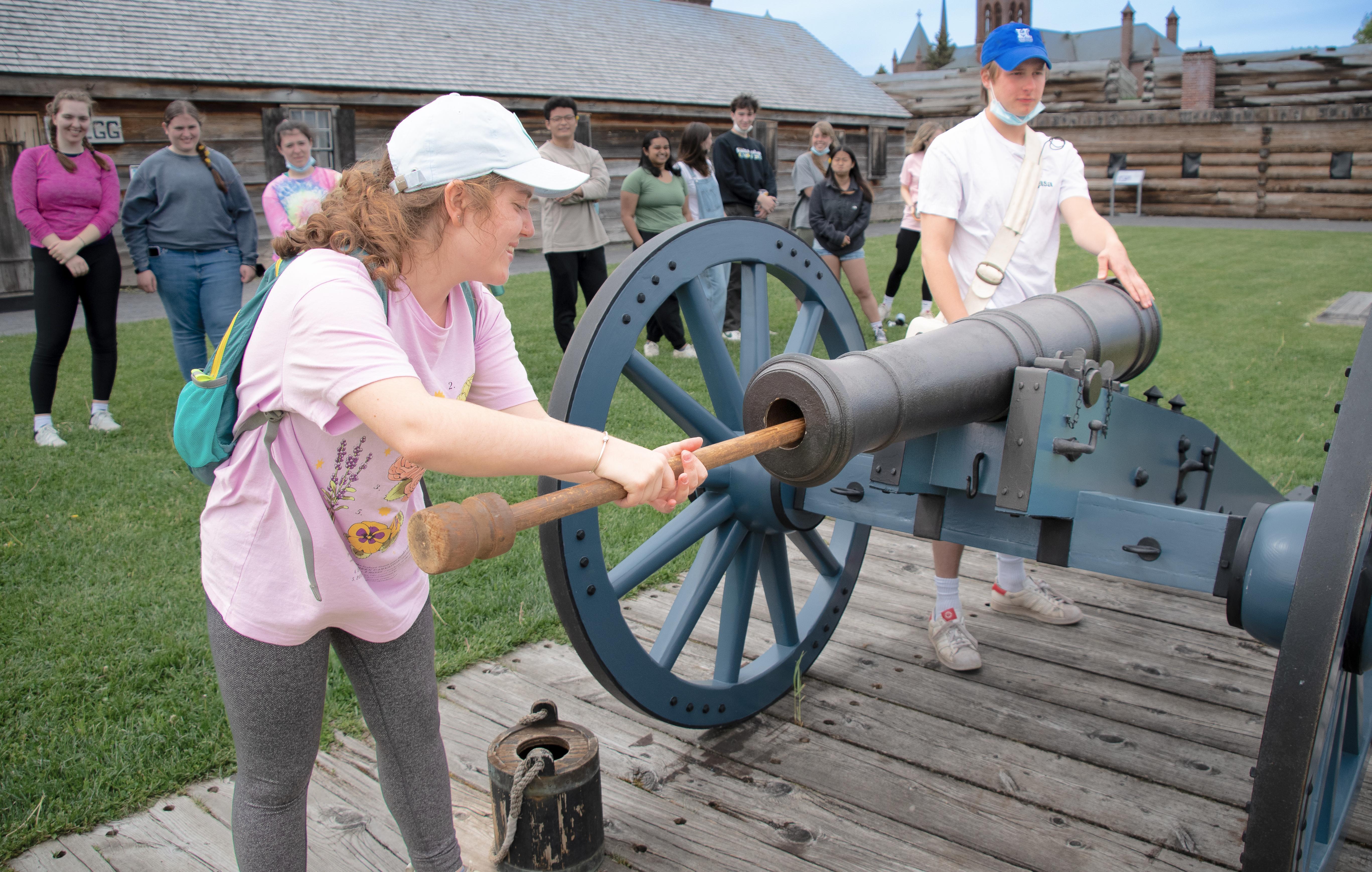 A young woman is using a long wooden q-tip like stick to clean a cannon while her friends watch on.