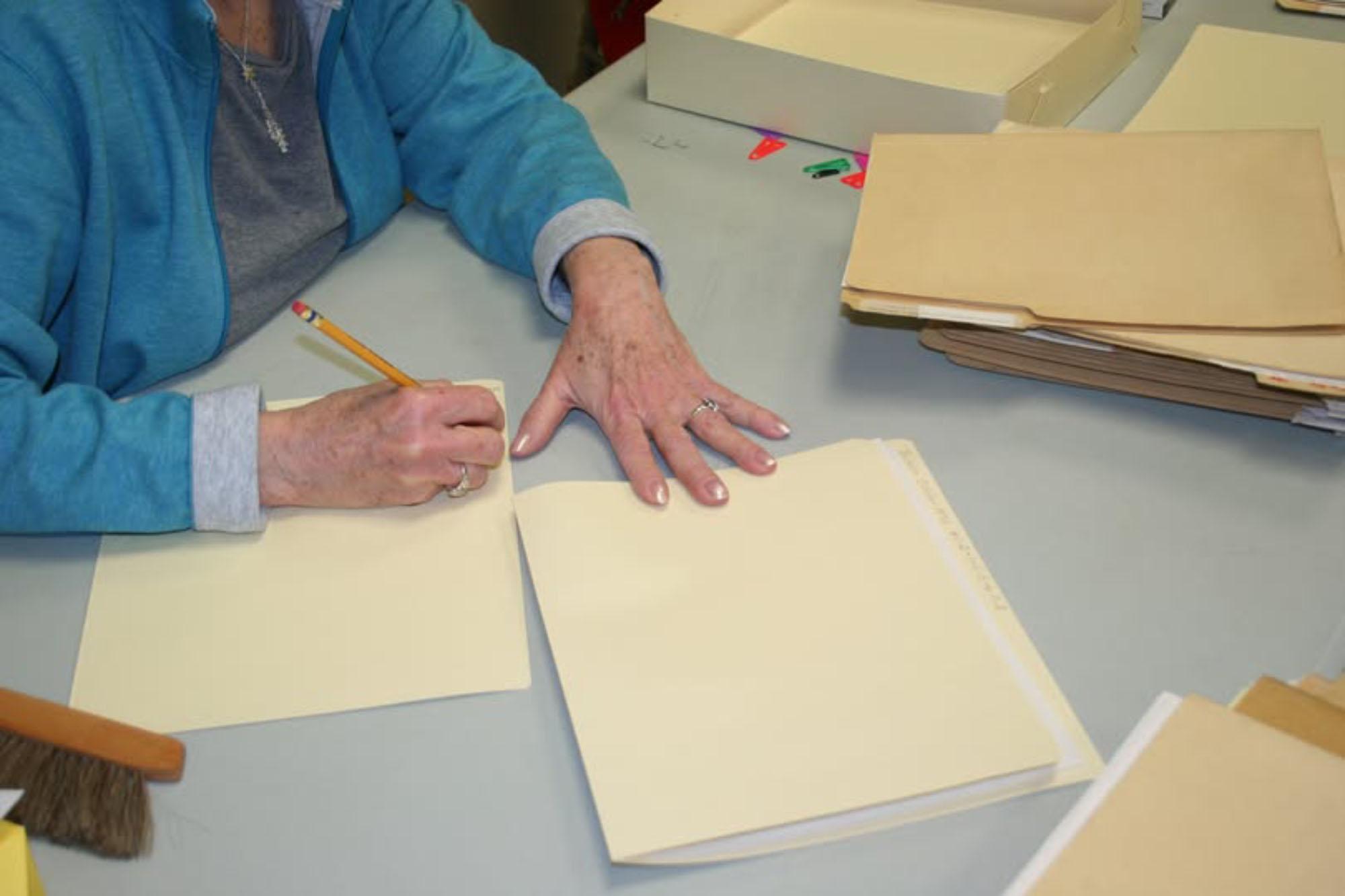 Camera view from above shows a table covered with folders. A cropped set of hands writes in pencil.