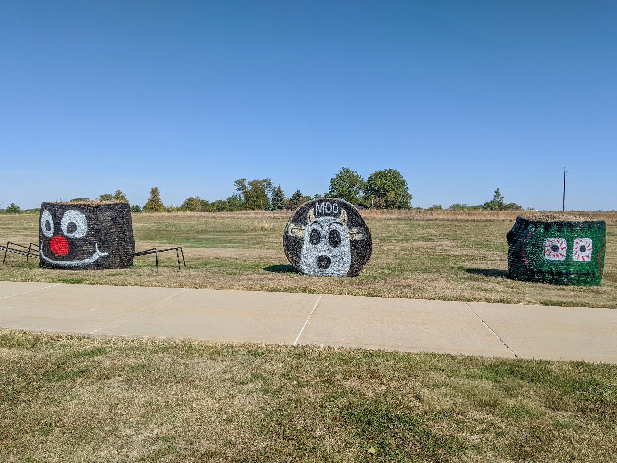3 painted hay bales (spider, cow, and monster) are seen in cut grass across from a paved walkway.