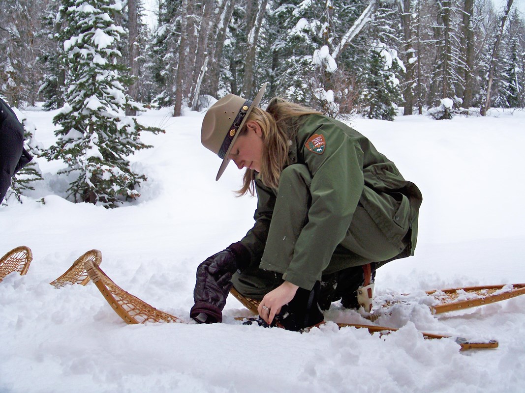 Ranger kneels in the snow to put on long wooden historic snowshoes.