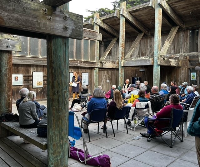 A group of people in a wooden courtyard, sitting in chairs looking at a person leading a song.