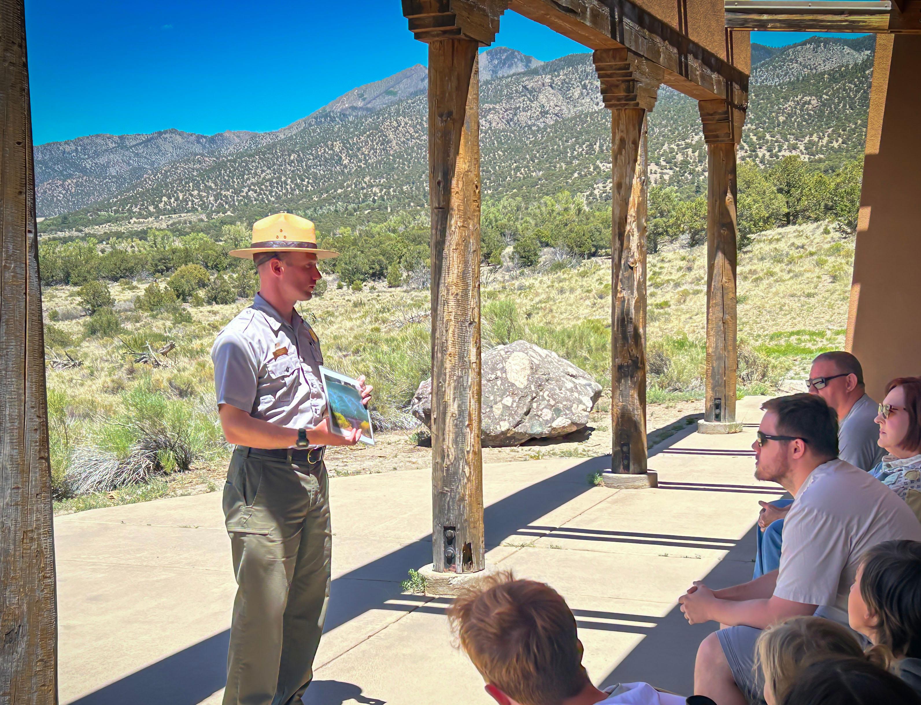 A park ranger in flat hat, grey shirt and green pants stands in front of visitors that are seated