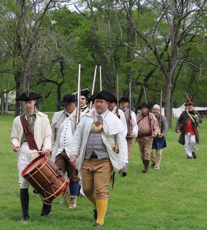 Men in colonial clothing marching behind a drummer.
