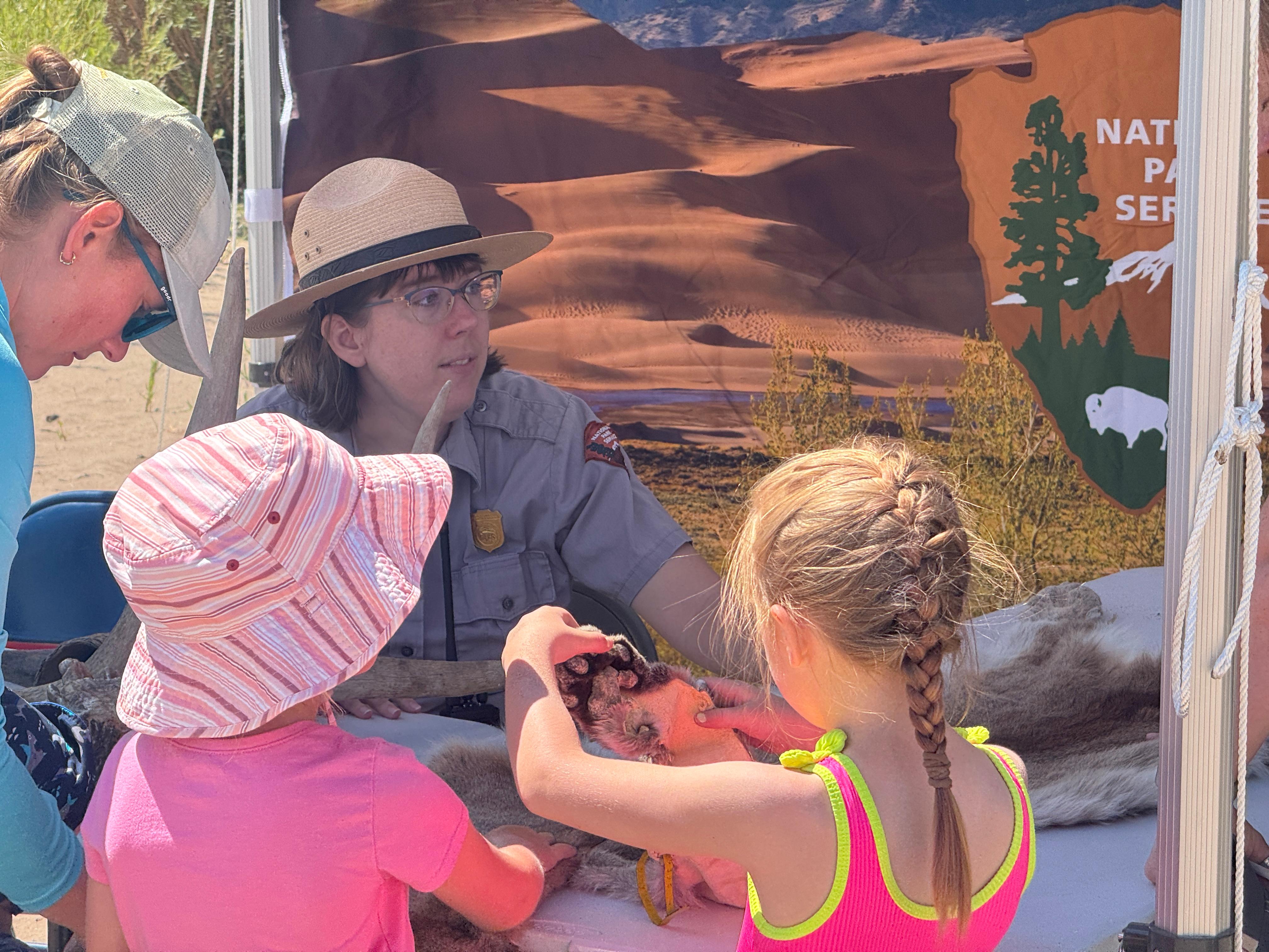 A park ranger in flat hat and grey uniform is facing two children and one at a table.