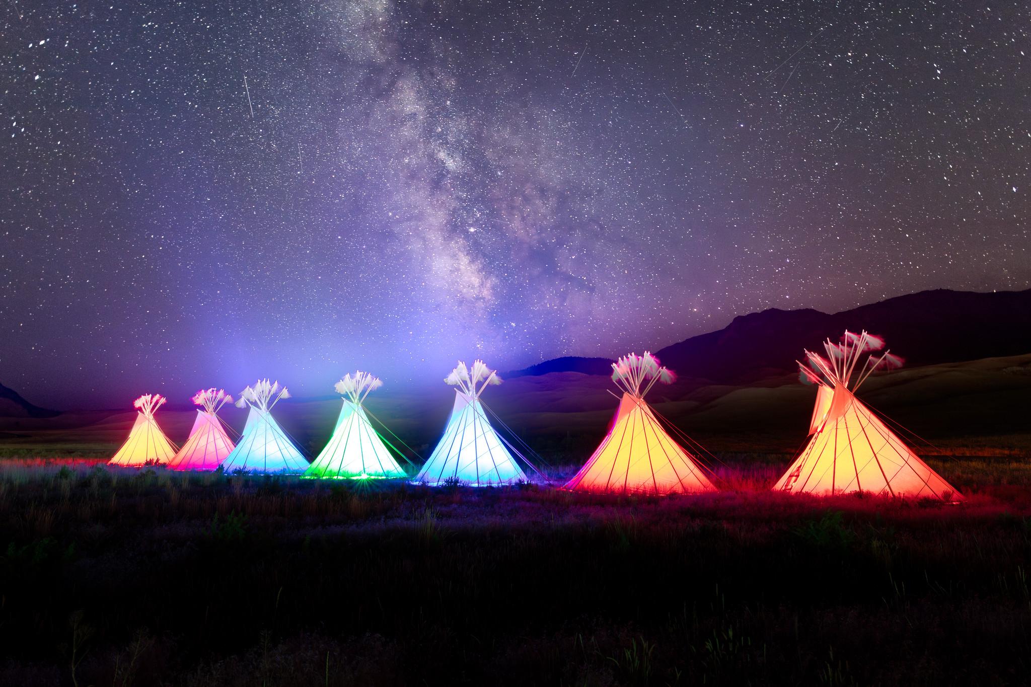 A row of illuminated, colorful teepees beneath a night sky filled with stars