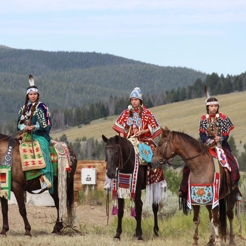 Three young women sit atop horses, all dressed in traditional regalia.