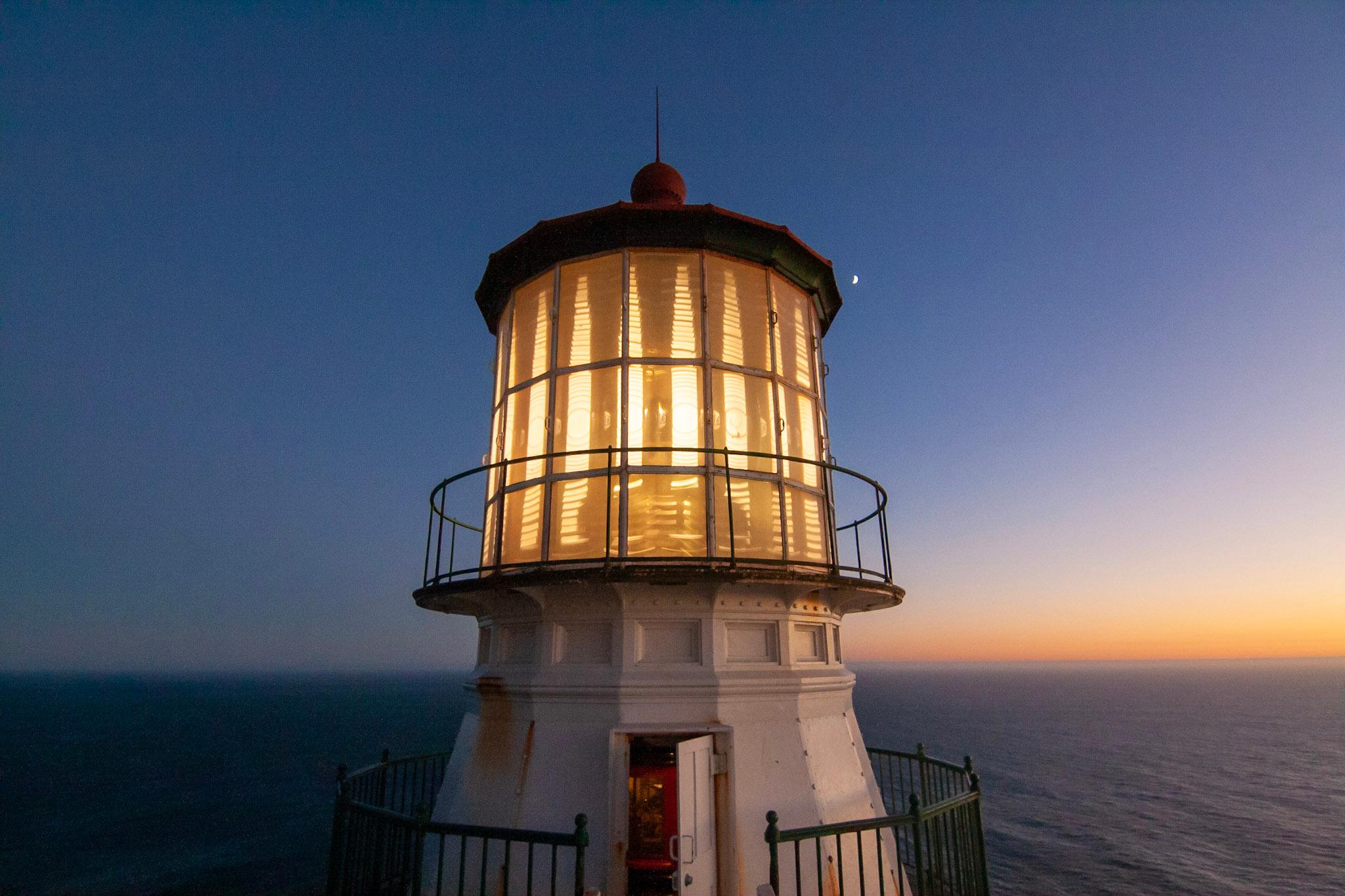 Light emanates from a short, white lighthouse in front of a dusky, sunset oceanscape.
