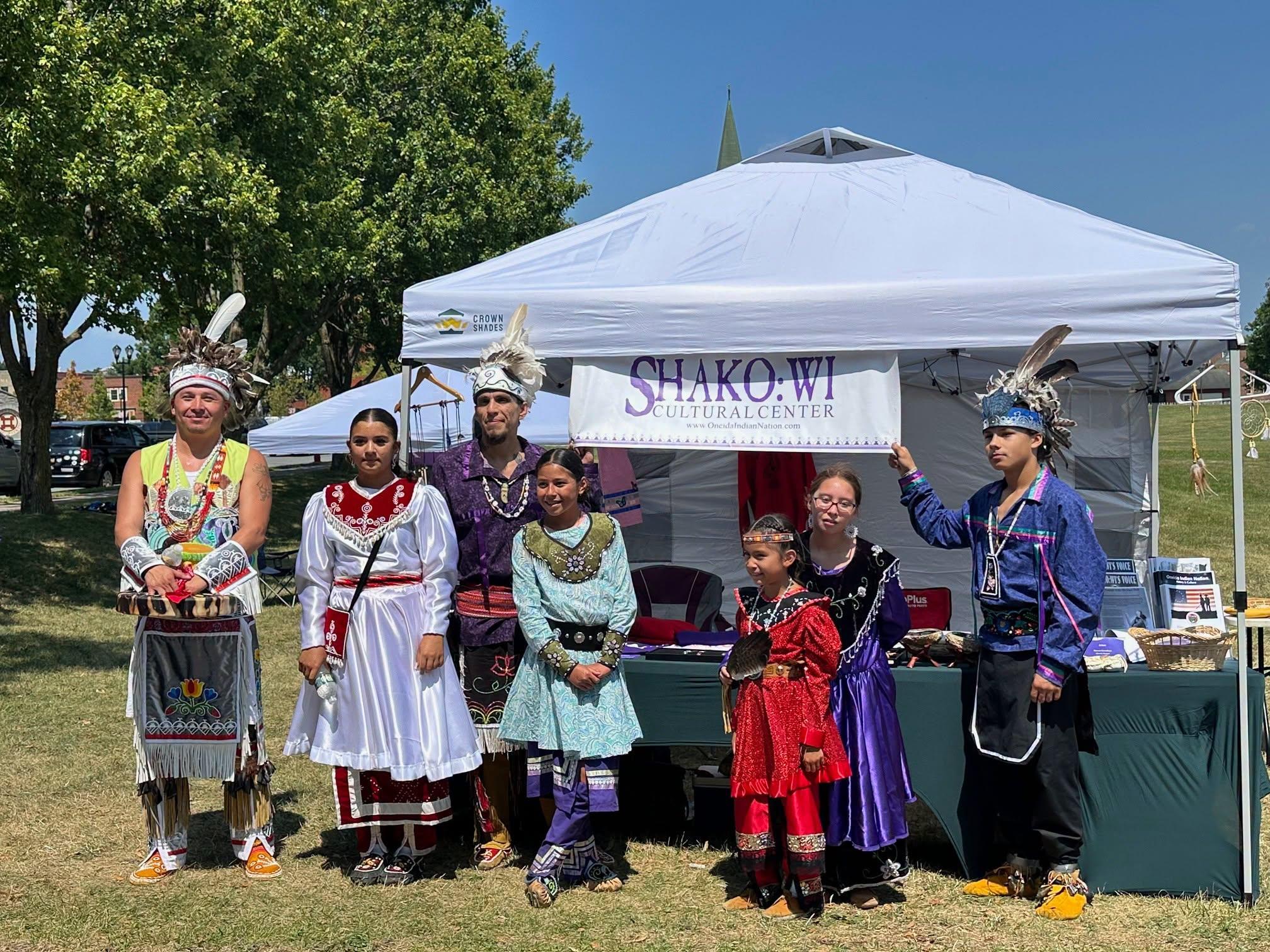 About a half dozen men women and children in traditional, colorful Oneida clothing pose and smile.
