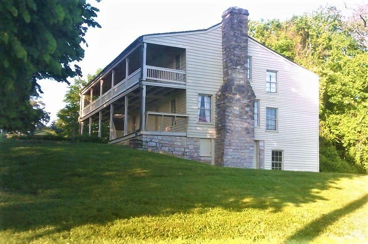 Historic two‑story wooden house with stone chimney on a grassy hill surrounded by trees.