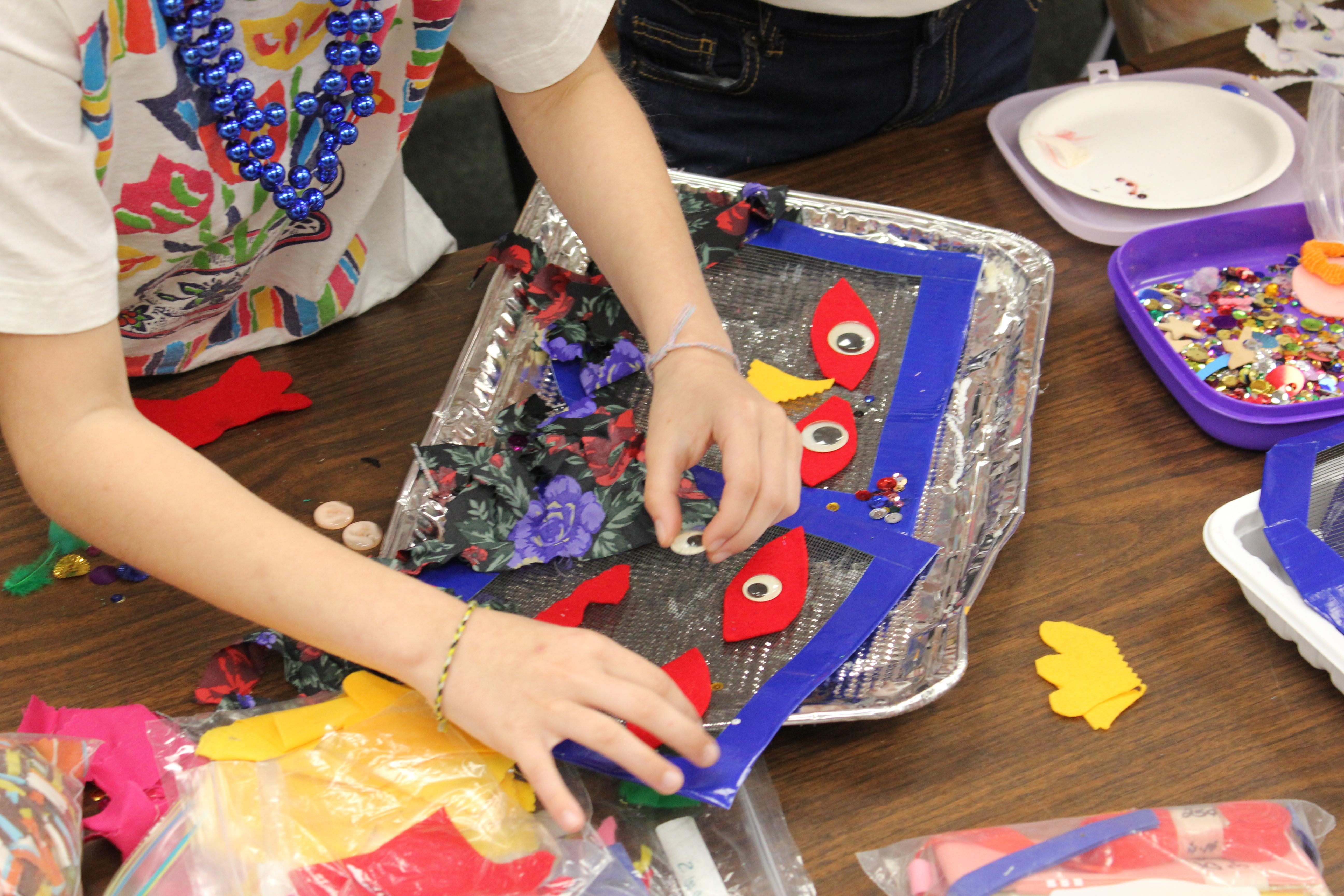 A child's hands add googly eyes to a mesh Mardi Gras mask.