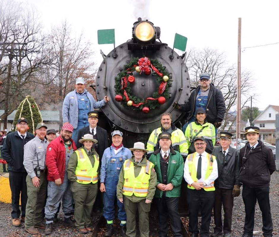 A group of 15 volunteers and employees pose in front of a steam engine decorated with a wreath