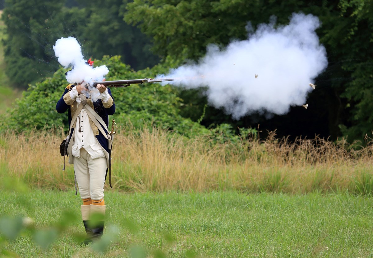 a continental army soldier in 18th century uniform fires a musket and the smoke covers his face