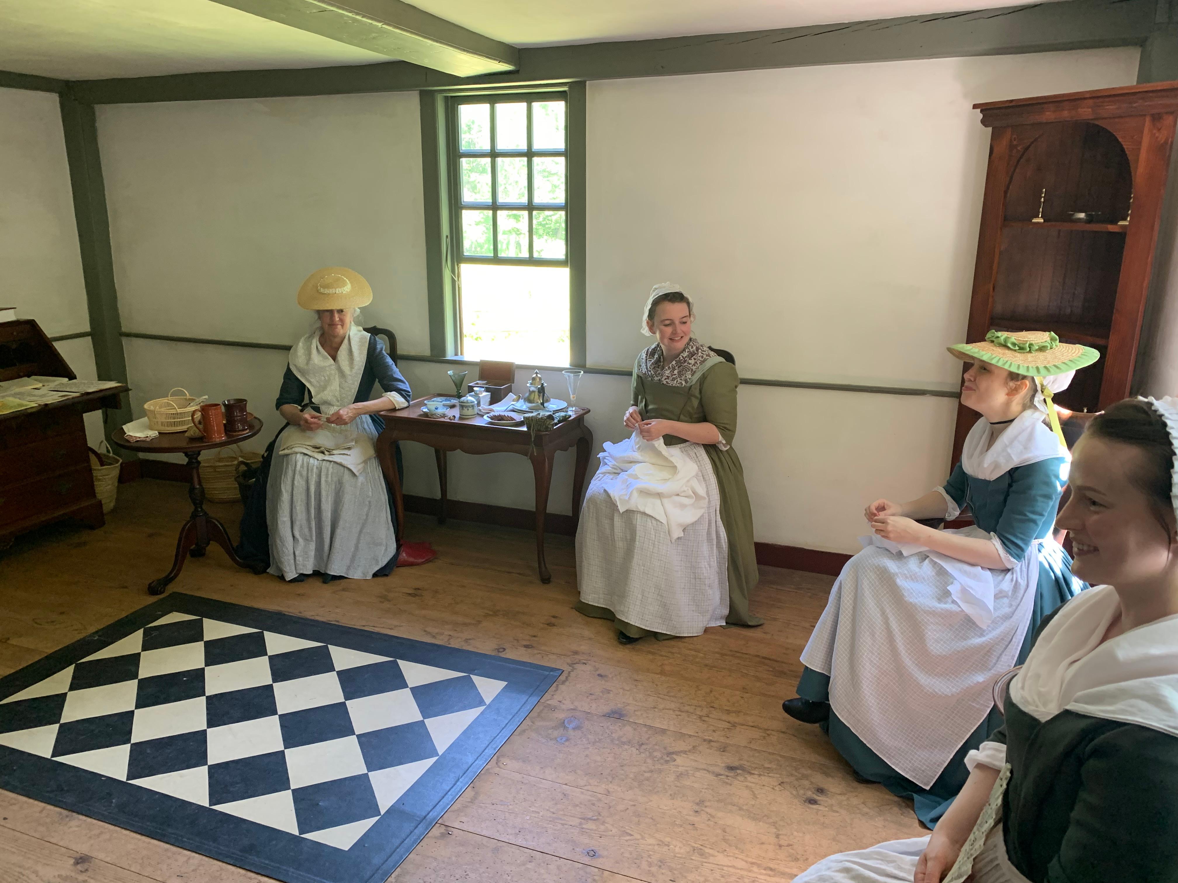 A group of colonial women sit in a parlor space talking