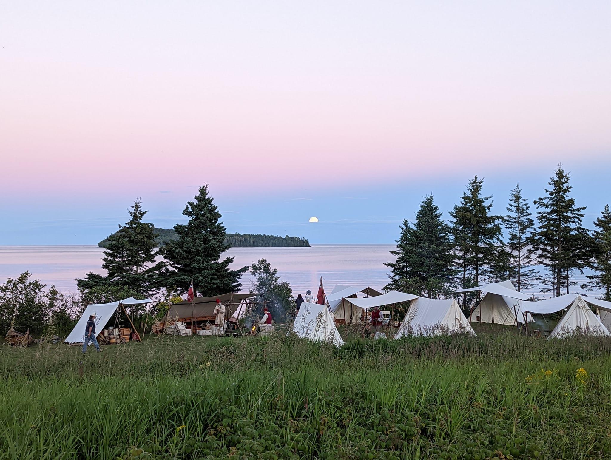 Moonrise over a bay with white canvas tents and costumed people in the foreground.